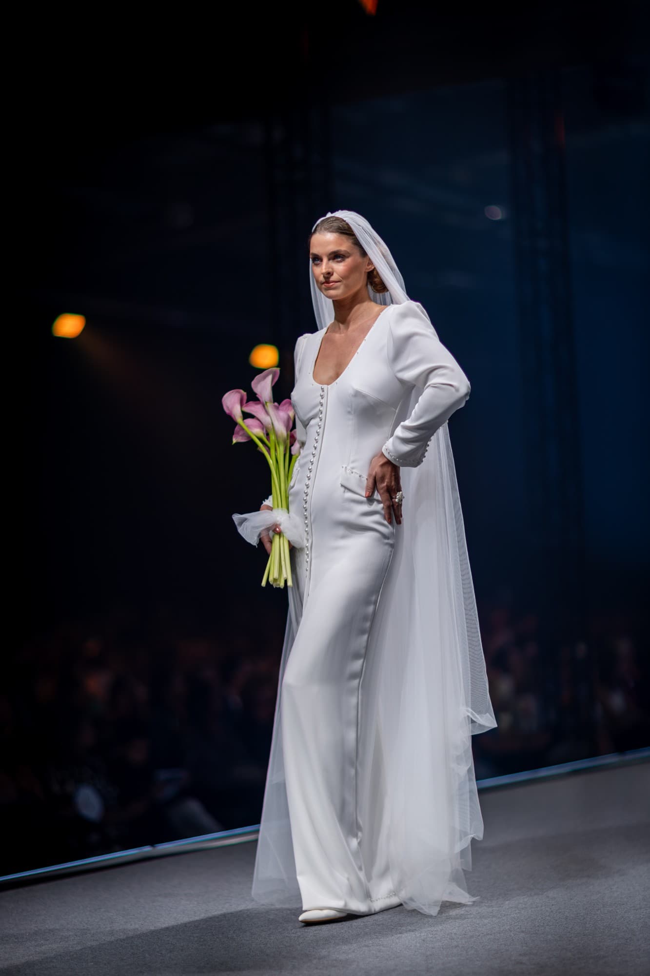 Mariée sur le podium en robe longue ivoire boutonnée de perles, voile de tulle et bouquet d’arums roses.